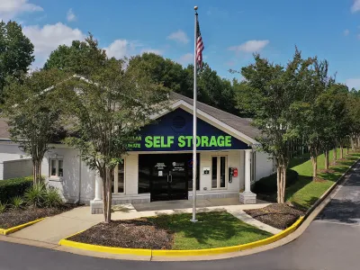 Self storage facility building with green signage, American flagpole, trees, and clear blue sky on a sunny day