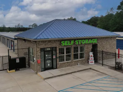 Brick and metal roof self storage office with green signage and multiple storage units behind it under cloudy sky.