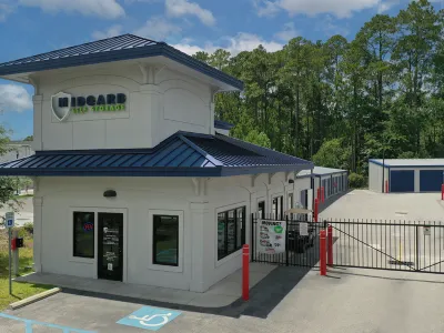 Midgard Self Storage facility with office building, gated entry, and storage units under blue sky and trees.