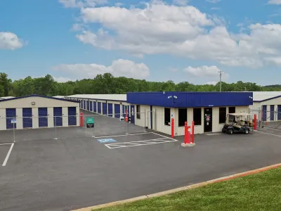 Self-storage facility with blue and white units, gated entrance, office building, and clear parking spaces under a partly cloudy sky.