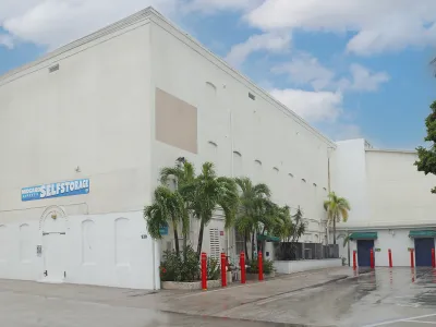 White self-storage building with palm trees and red bollards under a cloudy sky.