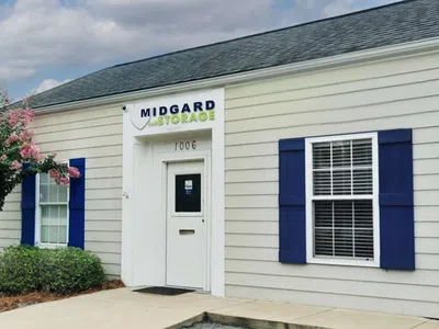 Self-storage facility with a gated entrance, small office building, and rows of storage units under a partly cloudy sky.