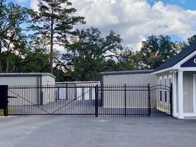 Gated entrance to a self-storage facility with storage units and an office building under a partly cloudy sky.