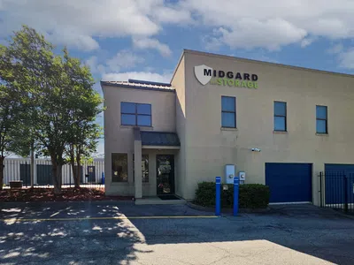 Modern beige Midgard Self Storage building with blue garage doors and trees under a partly cloudy sky.