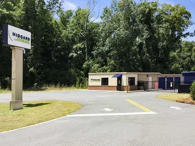 Midgard Self Storage facility with signage, office building, gated entrance, and surrounding greenery on a sunny day