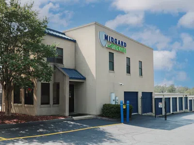 Modern beige Midgard Self Storage building with blue garage doors and trees under a partly cloudy sky.