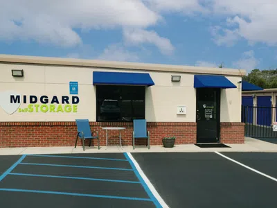 Midgard Self Storage facility with signage, office building, gated entrance, and surrounding greenery on a sunny day