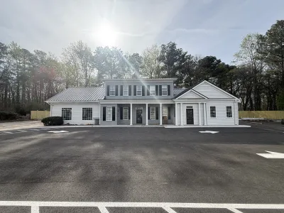 White two-story building with black shutters surrounded by trees and a large empty parking lot under a sunny sky