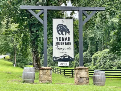 Wooden sign for Yonah Mountain Vineyards hangs between stone pillars with barrels at green forest roadside.