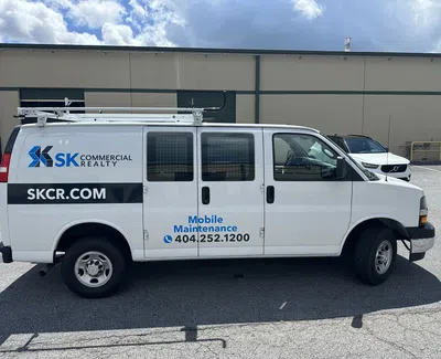 White SK Commercial Realty van with roof ladder rack and Mobile Maintenance contact info parked in lot under cloudy sky