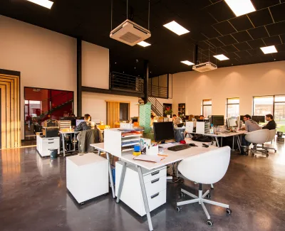 Open modern office space with multiple desks, computers, employees working, and large windows letting in natural light
