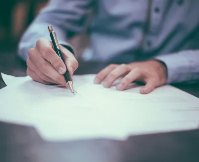 Person in a blue shirt writing on white paper with a pen at a desk in a blurred setting