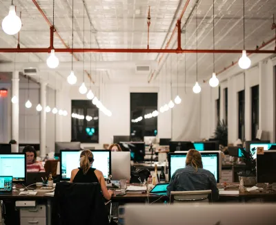 Modern office with people working late at computers under hanging light bulbs in open workspace at night.