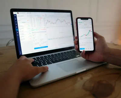 Person analyzing stock charts on smartphone and laptop with trading interface on wooden desk.
