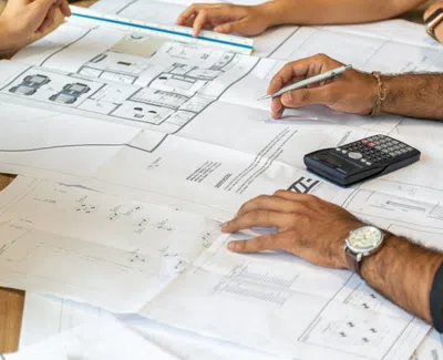 Two people working on architectural blueprints with a calculator and laptop on a wooden table.