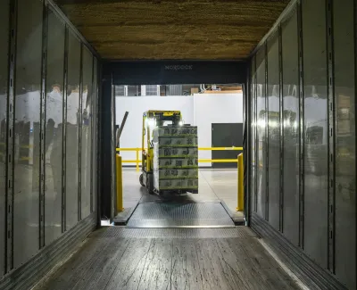 Forklift unloading stacked boxes from a truck at a warehouse loading dock with yellow safety rails