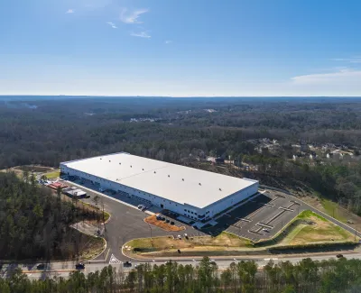 A large white industrial warehouse surrounded by forest and parking lots under a clear sky.