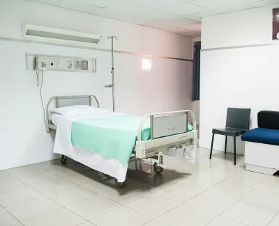 Clean hospital room with a single bed, teal blanket, black chair, and blue sofa against white tiled walls.