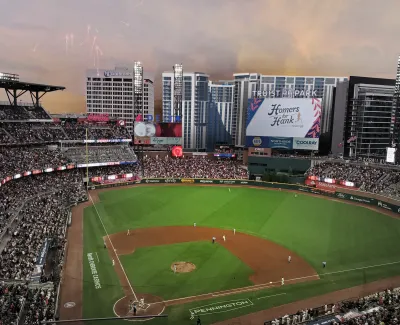 Aerial view of a crowded baseball stadium at dusk with fireworks and city buildings in the background
