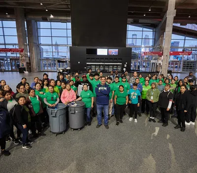 Large diverse group of volunteers in green shirts gathered indoors with trash bins for community cleanup event.