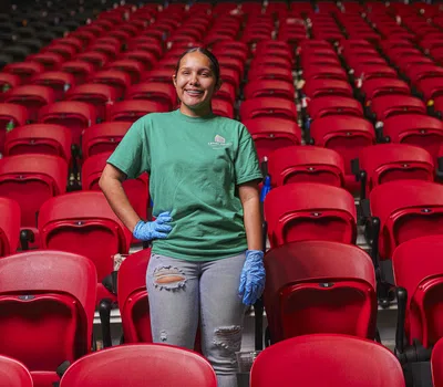 Smiling woman in green shirt and blue gloves standing among rows of empty red stadium seats.