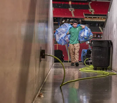 Person carrying bags of recyclables down a narrow stadium tunnel with cleaning equipment nearby