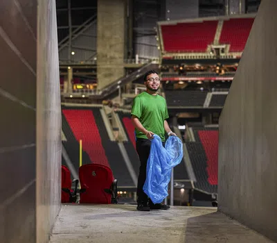 Man in green shirt holding a blue trash bag in an empty stadium corridor with red seats in the background