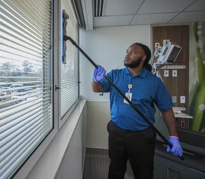 Janitor with gloves cleaning large hospital window blinds using a long-handled tool in a modern medical room.