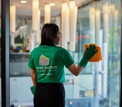 Woman wearing green shirt cleaning a large glass window with an orange cloth in a modern building.