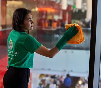 Professional cleaner in green gloves wiping a glass window with an orange cloth in an office building.