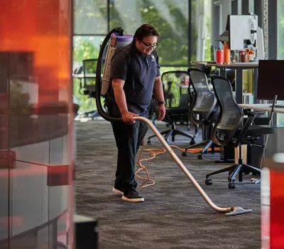 Professional cleaner vacuuming carpet in modern office space with desks, chairs, and large windows.