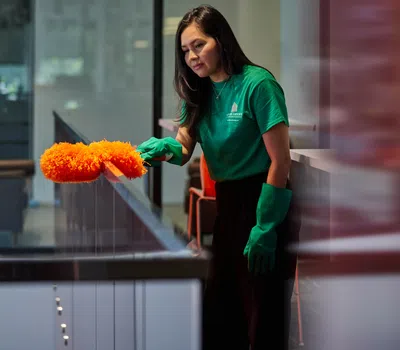 Woman in green shirt and gloves dusting glass railing indoors with orange duster in modern building.