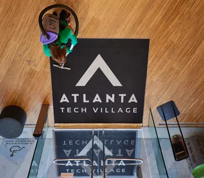 Person cleaning wooden floor near Atlanta Tech Village entrance with black logo mat and glass doors.
