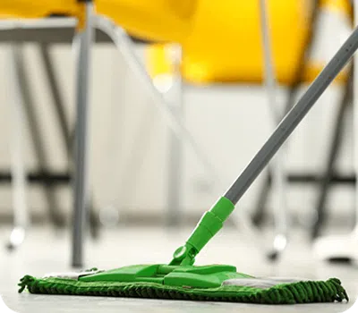 Green microfiber mop cleaning a light floor with yellow chairs blurred in the background in a bright room