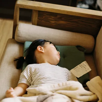 Young child sleeping on a beige couch with a blanket and pillow, resting peacefully indoors.