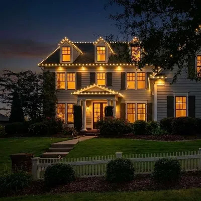 Large white house at dusk with warm interior lights and decorative string lights on the roofline, surrounded by greenery and a white fence.