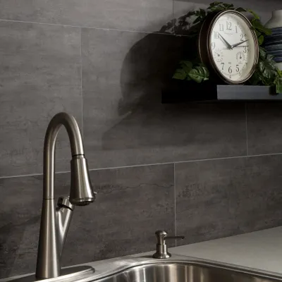 Modern stainless steel kitchen faucet with soap dispenser under clock on dark tiled backsplash.