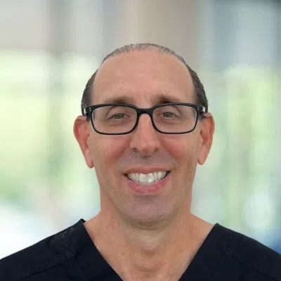 Smiling middle-aged man with glasses wearing black medical scrubs against a blurred background.