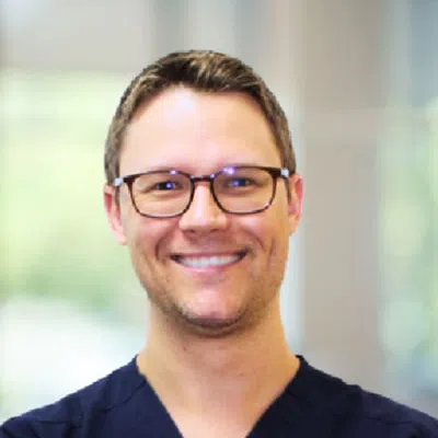 Smiling man wearing glasses and dark scrubs with blurred indoor background.