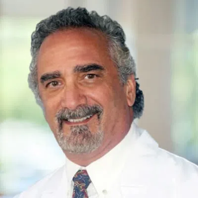 Smiling middle-aged male doctor with gray hair and beard wearing a white coat and patterned tie indoors.