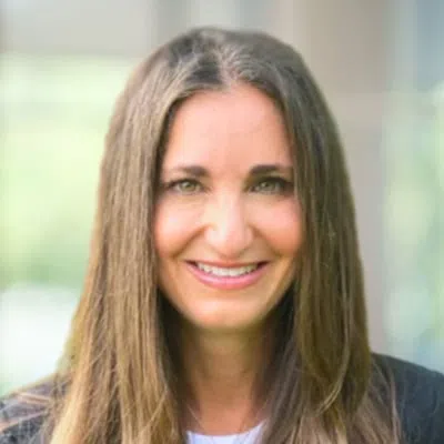Smiling woman with long straight brown hair and green eyes wearing a black jacket and white top outdoors.
