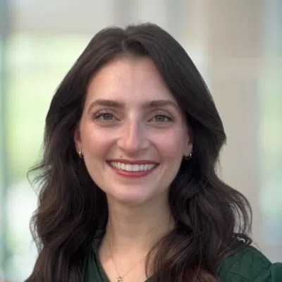 Smiling woman with long dark hair wearing a green top and gold necklace in a bright indoor setting