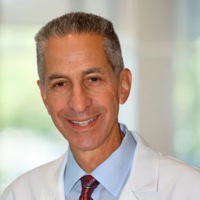 Smiling middle-aged male doctor wearing a white coat, blue shirt, and red paisley tie in a bright office setting.