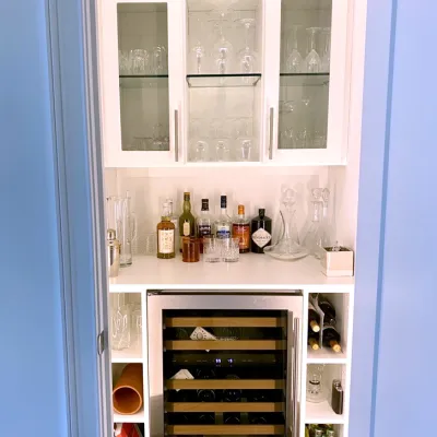Home wine bar with white shelves, glass doors, wine bottles, liquor bottles, wine fridge and glassware