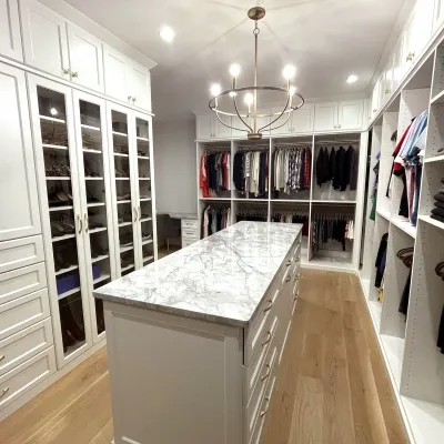 Spacious walk-in closet with marble island, white cabinets, white shelves, glass doors, hanging clothes, shoes, brushed nickel light fixture and hardwood floor
