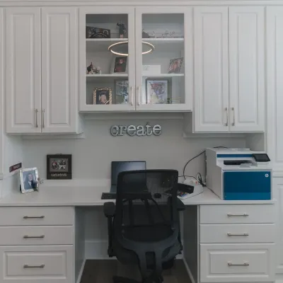 Modern home office with white cabinets, glass cabinet doors, white desk drawers, laptop, printer, black executive chair and decorative create sign on wall