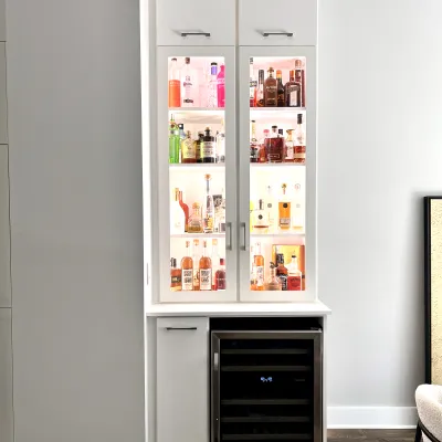 Home bar with white built-in cabinets with glass doors displaying a variety of liquor bottles, a mini fridge and accent lighting