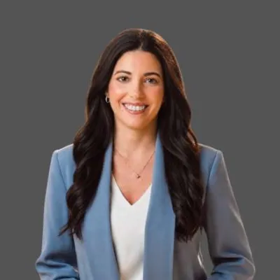 Smiling woman with long dark hair wearing a light blue blazer and white top on a gray background