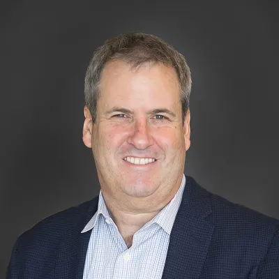 Smiling middle-aged man in a dark blazer and checkered shirt against a solid dark background