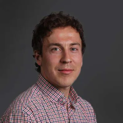 Portrait of a young man with curly brown hair wearing a checkered shirt against a gray background